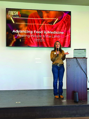 A woman stands at a podium, speaking in front of a screen displaying the presentation title "Advancing Food is Medicine: Healing People & the Land" at a FreshRx Oklahoma event.