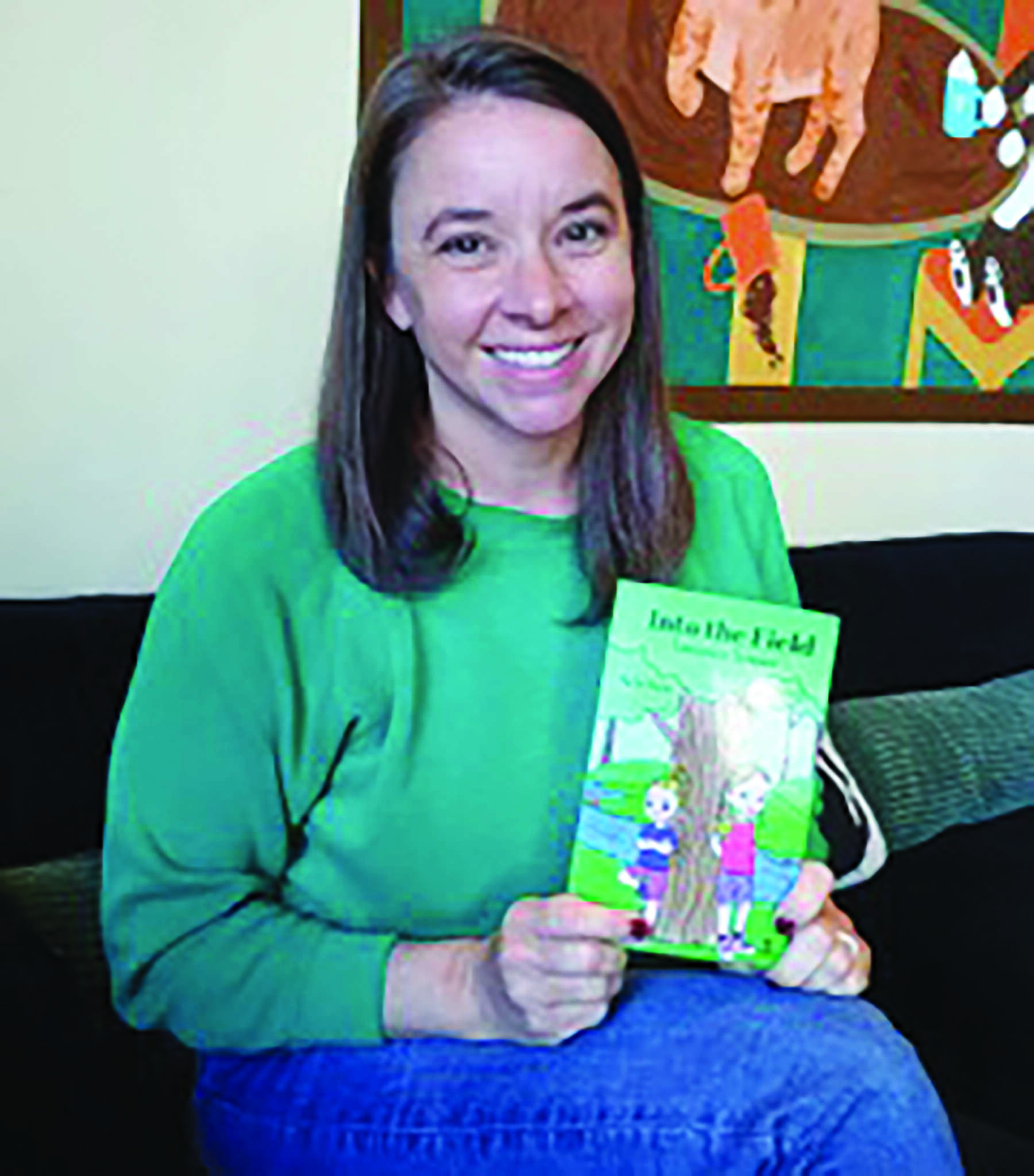 A woman in a green sweater sits on a couch, smiling and holding a children's book titled "Into the Field." A colorful painting hangs on the wall behind her.
