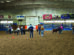 People and children with ponies or miniature horses stand in a dirt-floor indoor arena, participating in a livestock or equestrian competition.