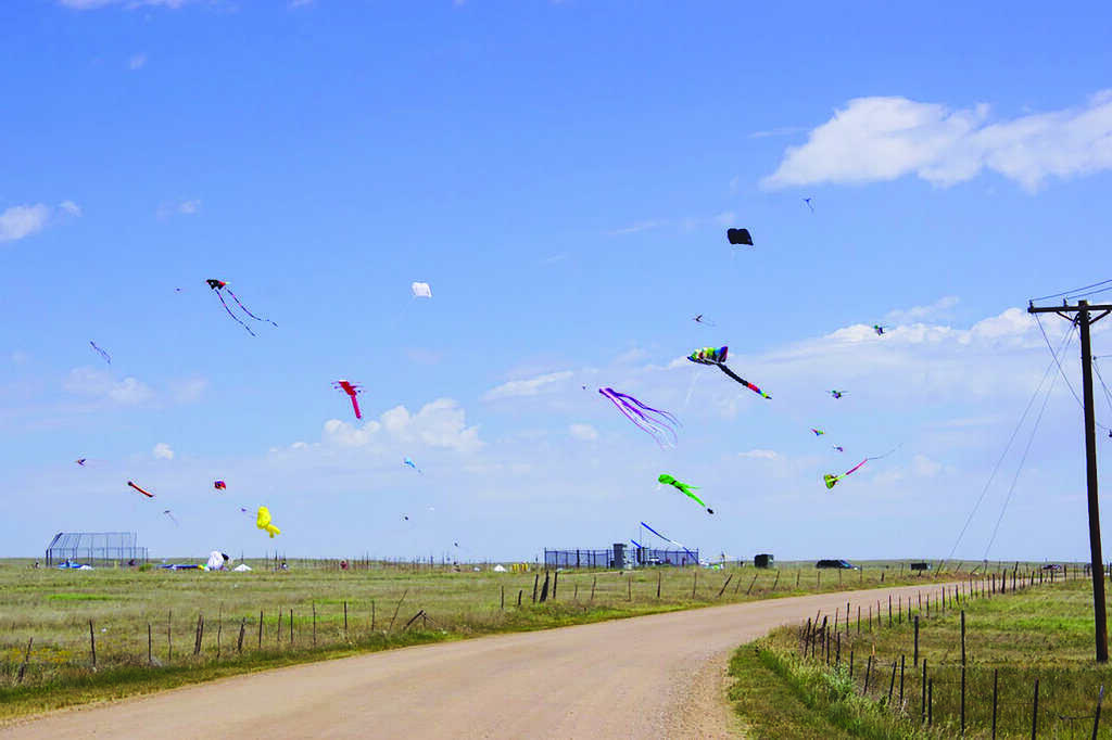 Several colorful kites fly in the blue sky above a rural landscape with a dirt road, grassy fields, and distant structures under scattered clouds.