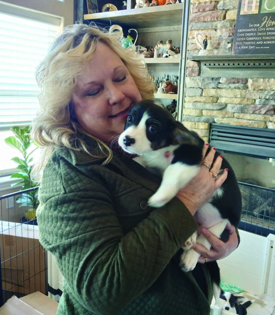 Woman in a green jacket holding a black and white puppy indoors, standing in front of a fireplace with various decorations on the mantel.