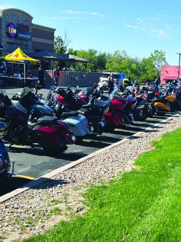 A row of motorcycles is parked along the curb outside a restaurant on a sunny day, with a grassy area and people visible in the background.