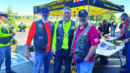 Three men in biker vests stand together and smile in front of an American Legion tent at an outdoor event, with other people and motorcycles in the background.