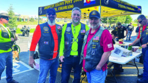 Three men in biker vests stand together and smile in front of an American Legion tent at an outdoor event, with other people and motorcycles in the background.