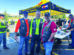 Three men in biker vests stand together and smile in front of an American Legion tent at an outdoor event, with other people and motorcycles in the background.