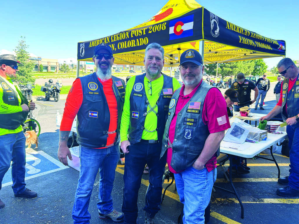 Three men in biker vests stand together and smile in front of an American Legion tent at an outdoor event, with other people and motorcycles in the background.