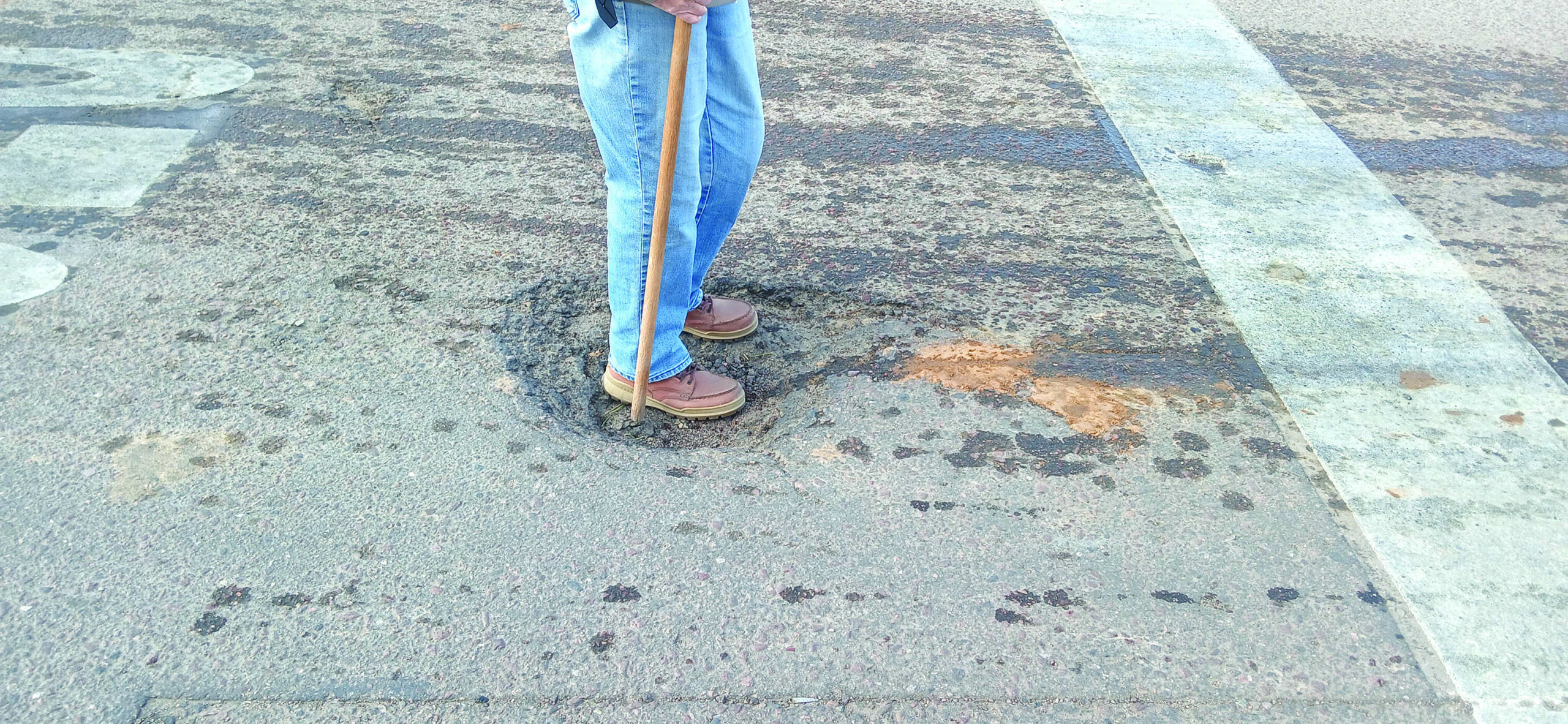 Person standing in a pothole on a road, holding a stick, with a crosswalk visible nearby.