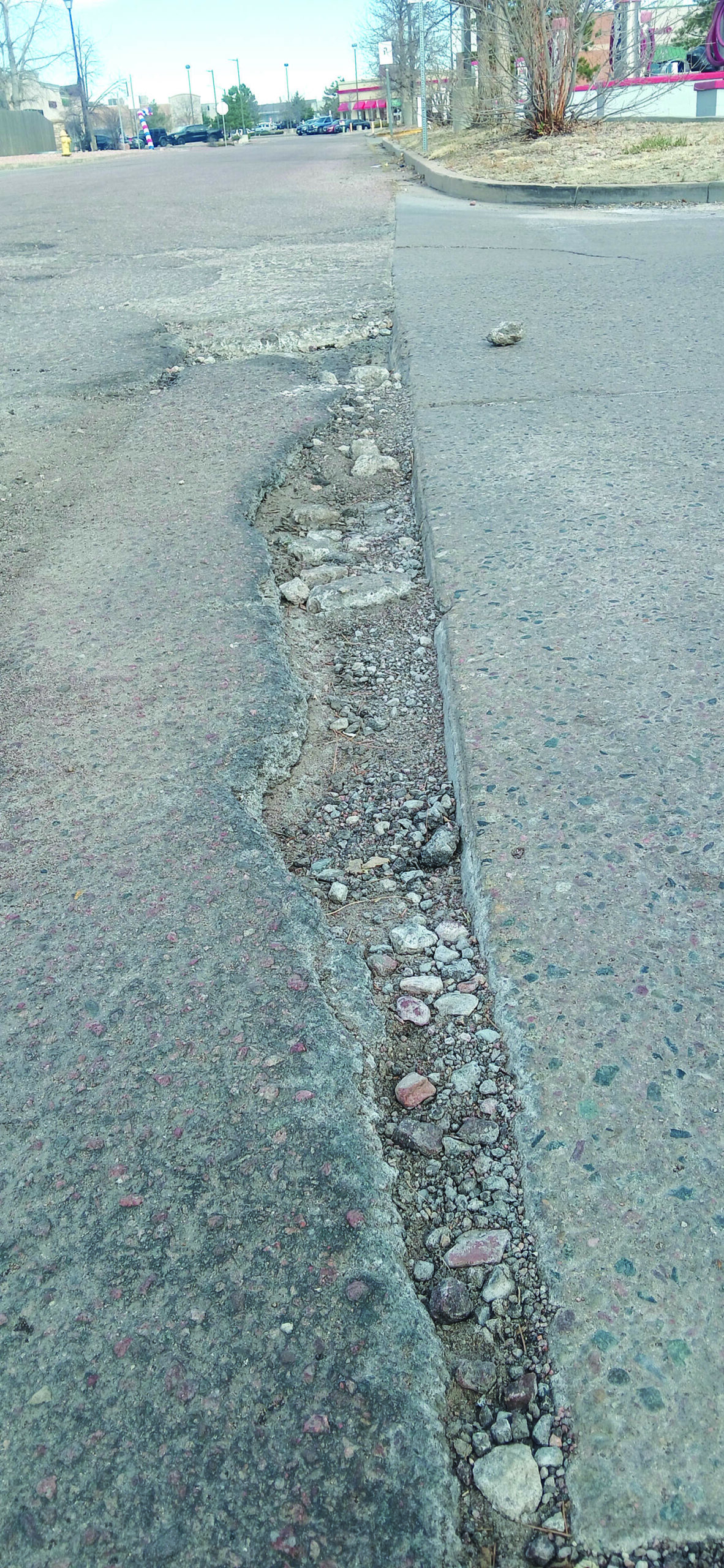 A close-up view of a cracked and uneven road surface, showing exposed aggregate and significant pavement damage along the edge.