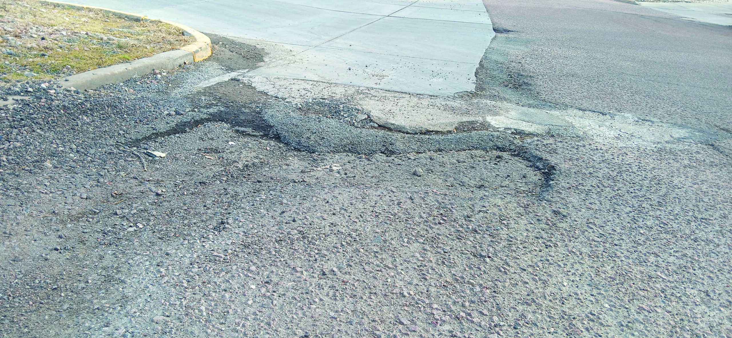 Damaged road surface with cracks and loose gravel near a concrete curb at a street corner.