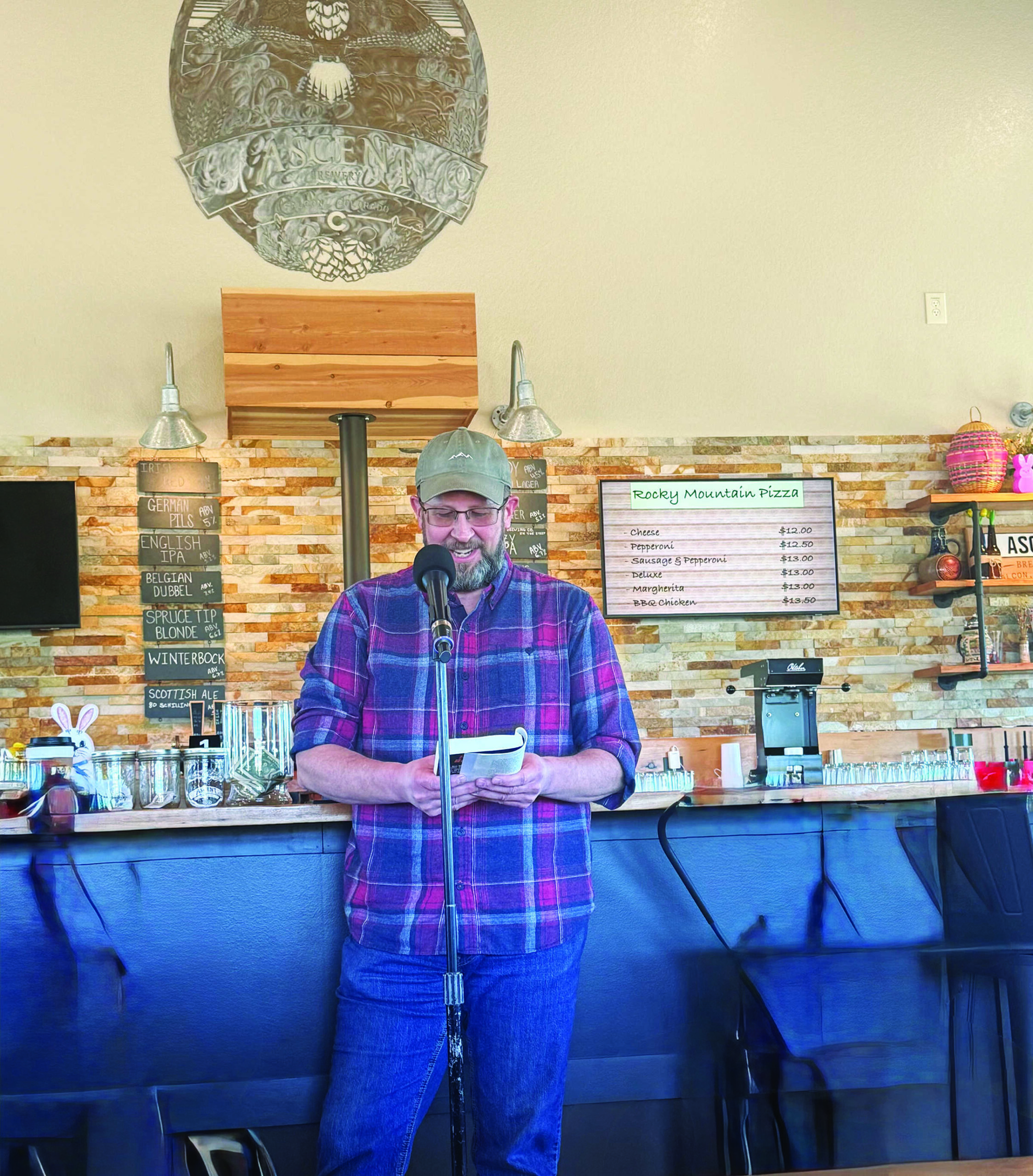 A man in a plaid shirt and cap stands at a microphone reading from a small notebook in front of a bar with menus and bottles in the background.