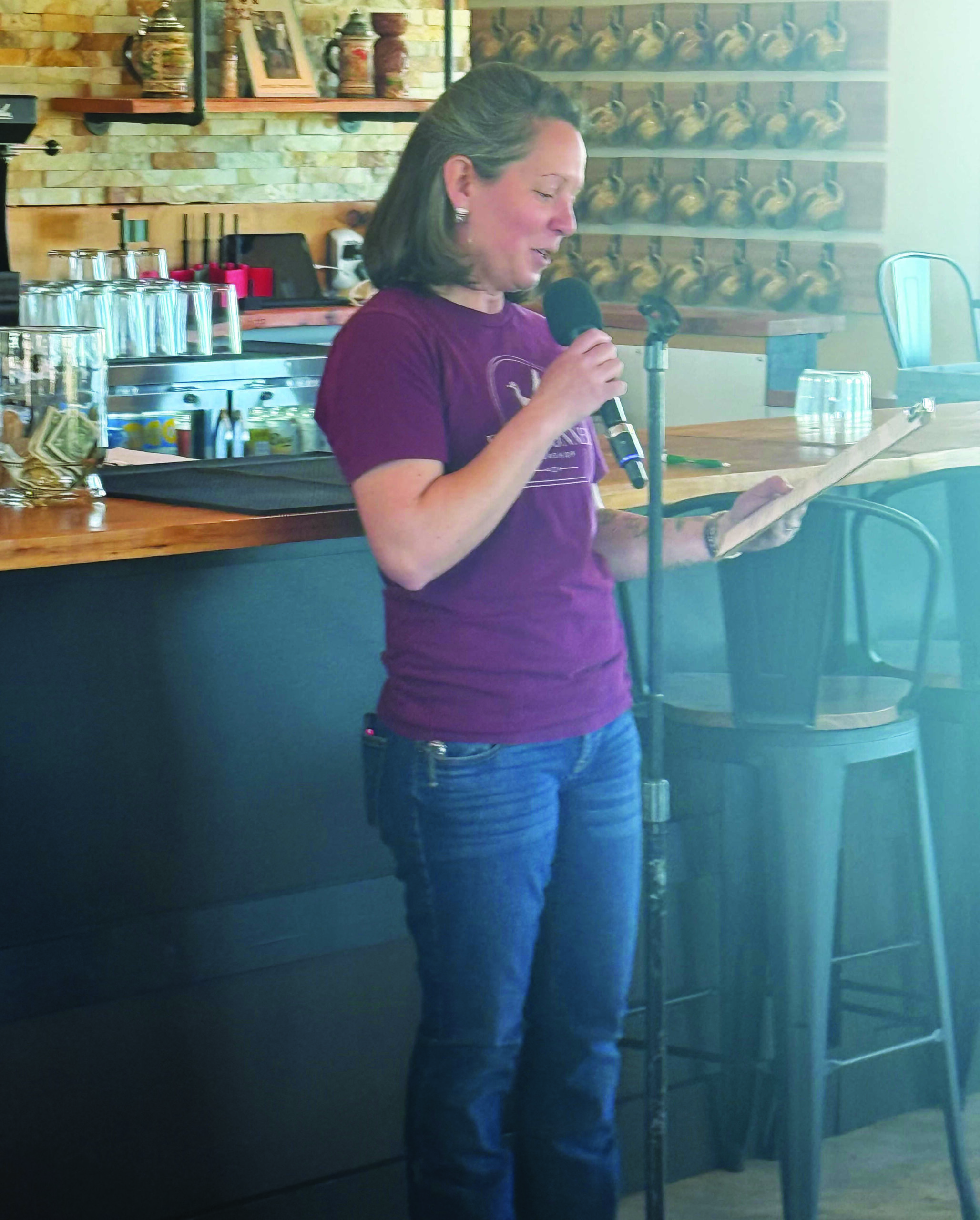 A woman stands at a microphone reading from a clipboard in front of a bar with stools, glassware, and various decor in the background.