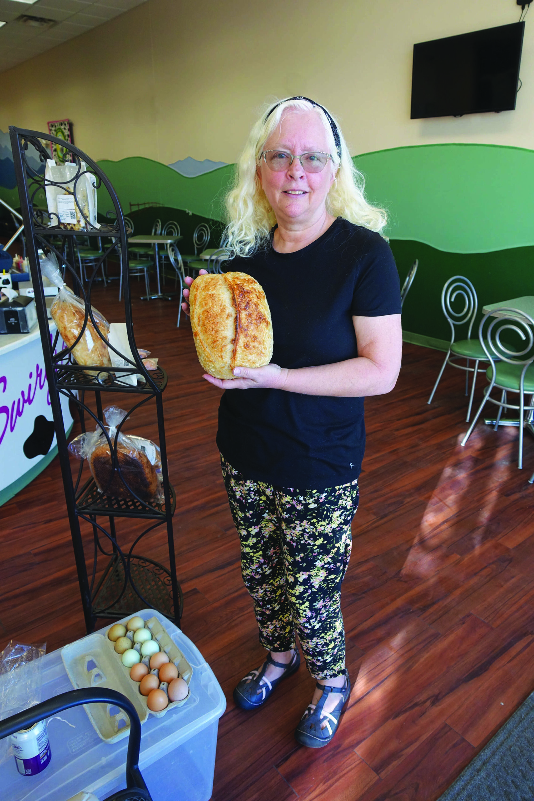 A woman with glasses and blond hair stands indoors, holding a loaf of bread. Eggs are on a plastic container nearby. The room has green and beige walls, wood flooring, and empty chairs.