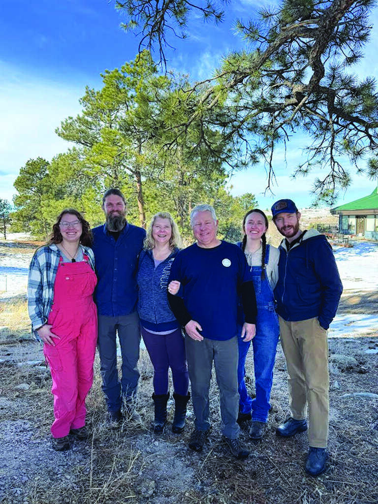 Six people stand together outdoors on a partly snowy day, with trees and a house in the background. They are smiling and dressed in casual winter clothing.