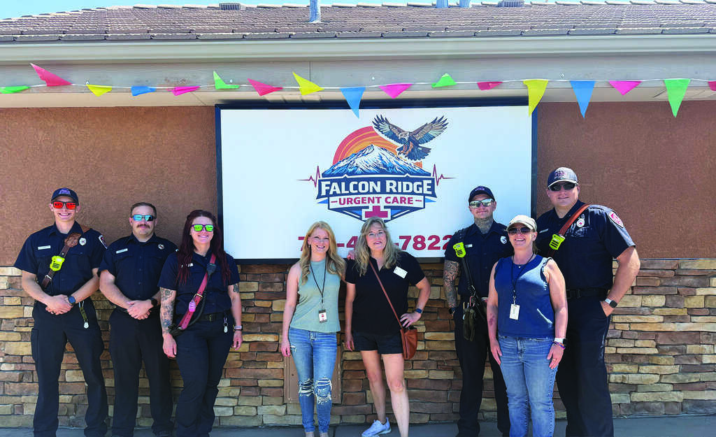 A group of eight people, including uniformed emergency personnel and civilians, stand in front of a Falcon Ridge Urgent Care sign with colorful pennant banners above them.