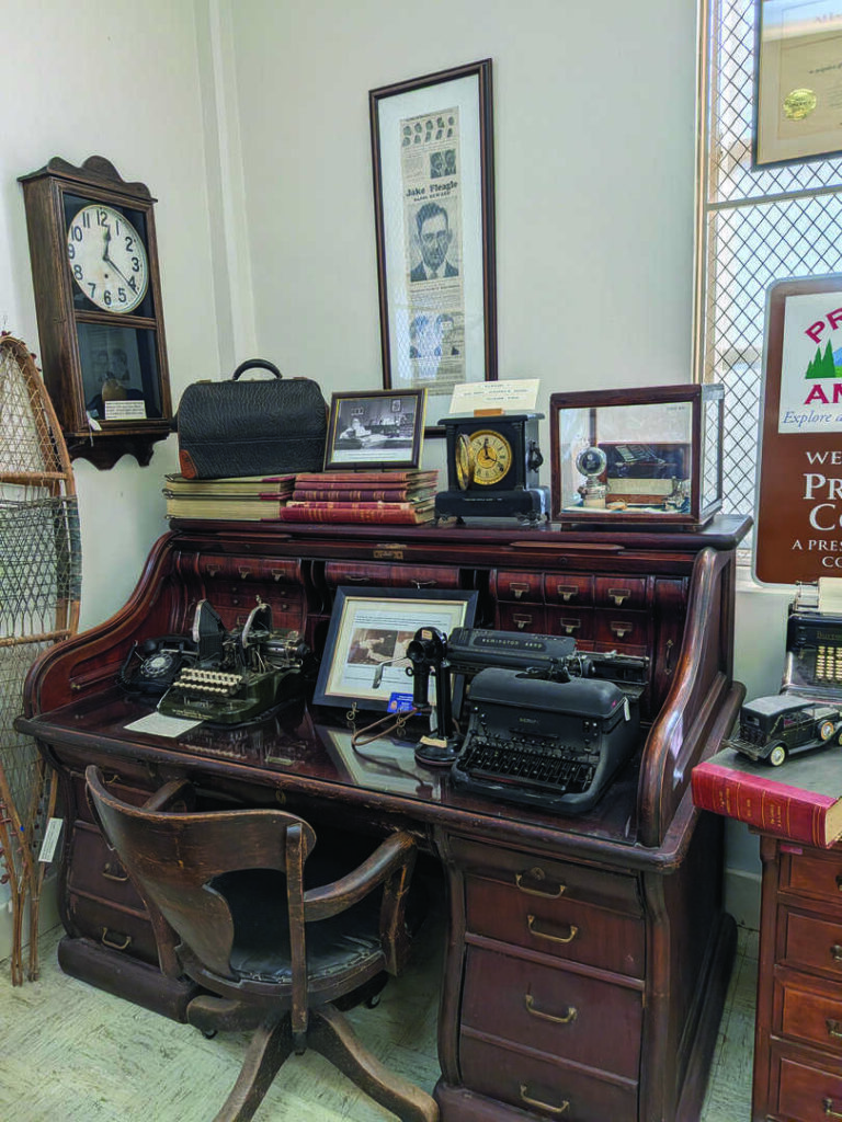 Antique rolltop desk with typewriter, telephone, old books, photos, and clocks. Wooden chair in front. Vintage decor and signs on the wall. Natural light from window at right.