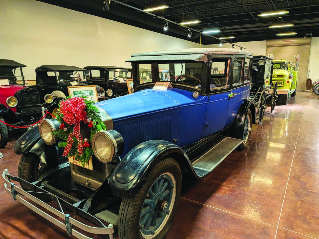 A vintage blue car with a wreath and red bow on the front is displayed indoors with several other classic cars in the background.