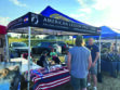 People stand and talk at an American Legion Post 2008 booth with a banner, brochures, and a table covered by a US flag at an outdoor event.