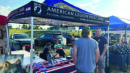 People stand and talk at an American Legion Post 2008 booth with a banner, brochures, and a table covered by a US flag at an outdoor event.