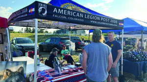 People stand and talk at an American Legion Post 2008 booth with a banner, brochures, and a table covered by a US flag at an outdoor event.