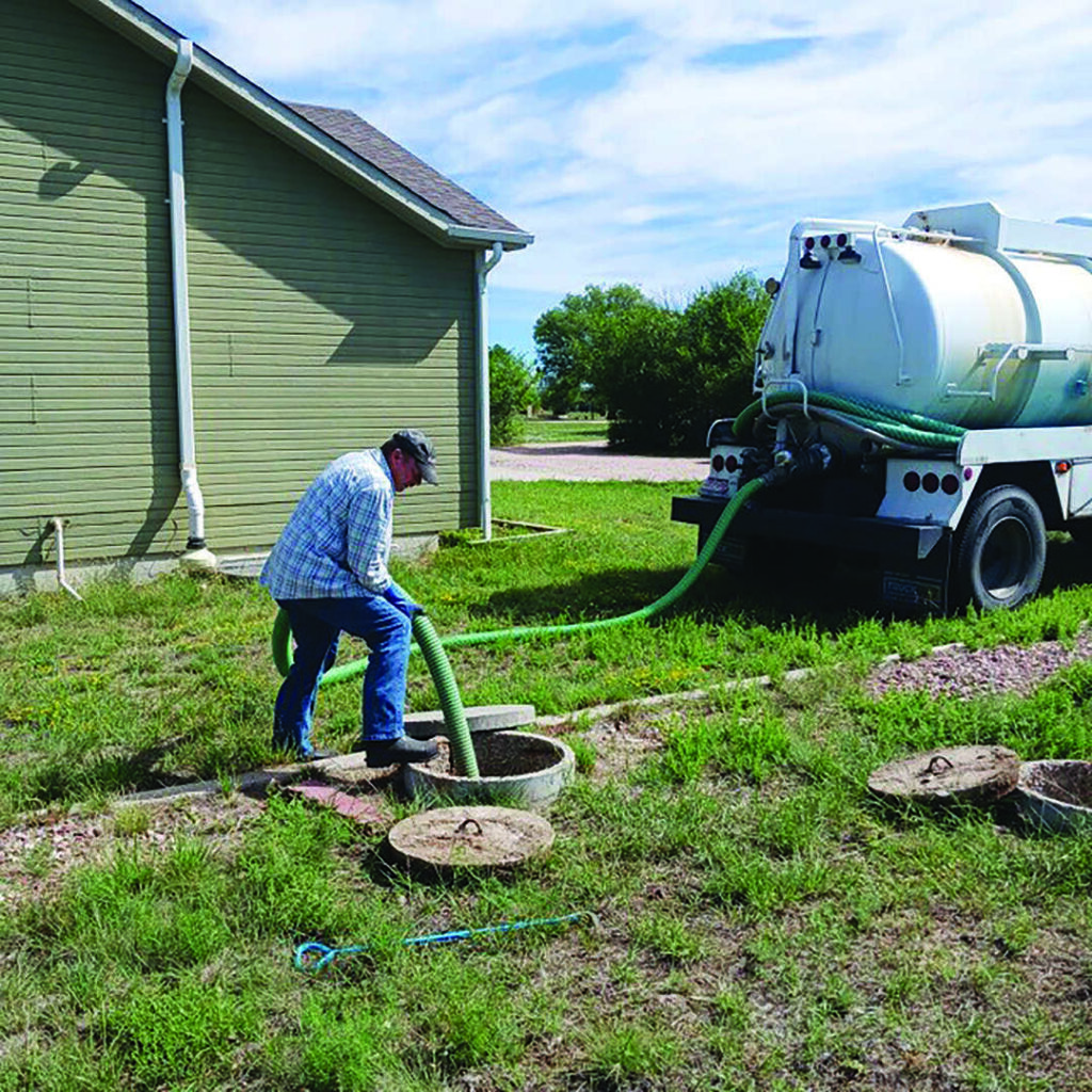 A person is connecting a green hose from a white septic truck to an underground tank beside a green building.