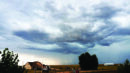 Dark storm clouds gather over a rural landscape with a house and fields.