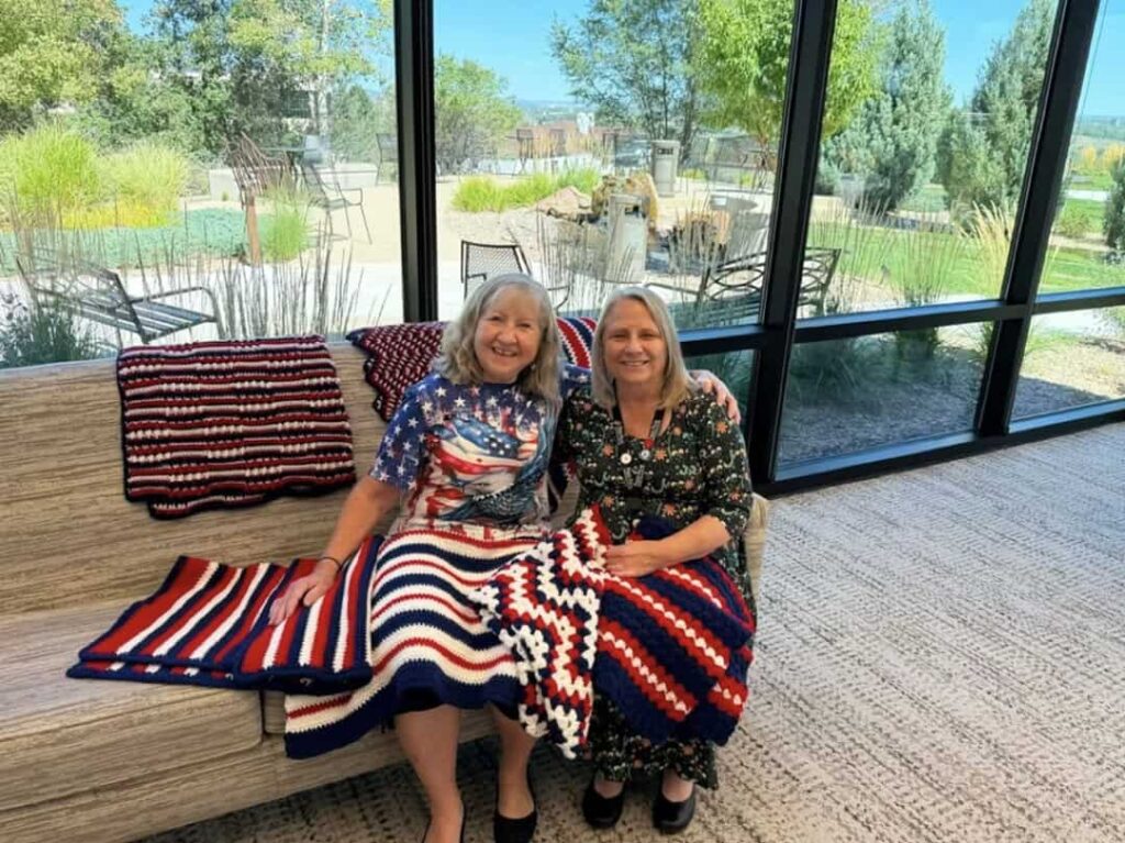 Two women sit on a sofa in front of large windows, displaying three handmade red, white, and blue crochet blankets.