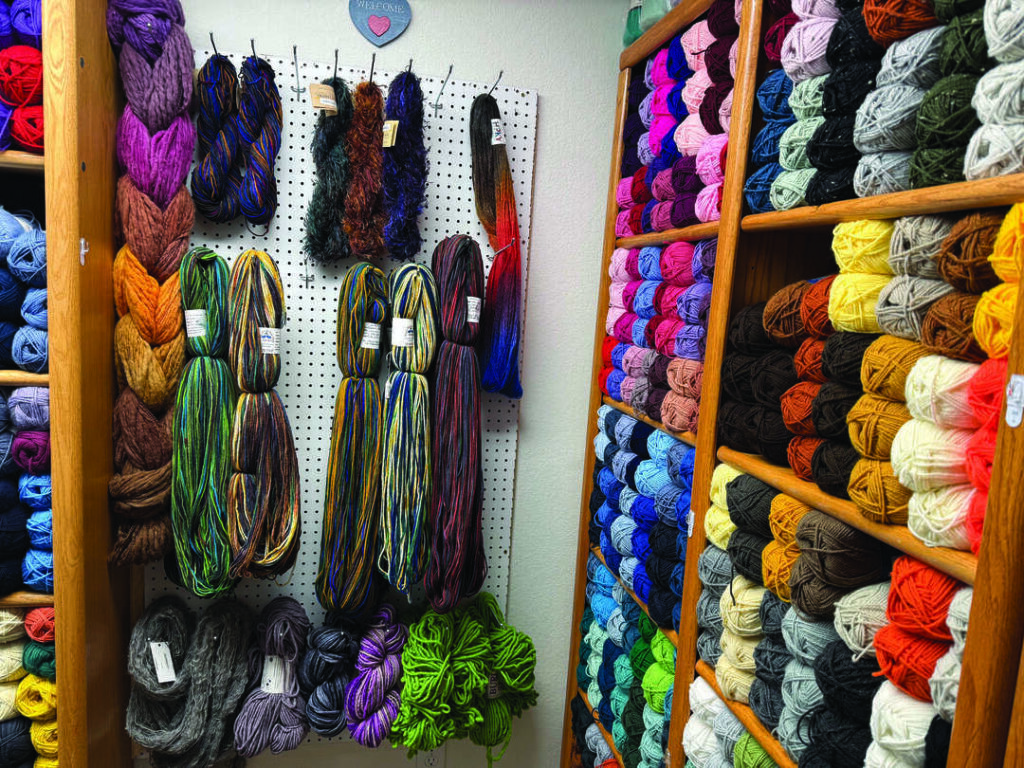 Skeins of multicolored yarn hanging on a wall pegboard beside wooden shelves stocked with stacked yarn balls.